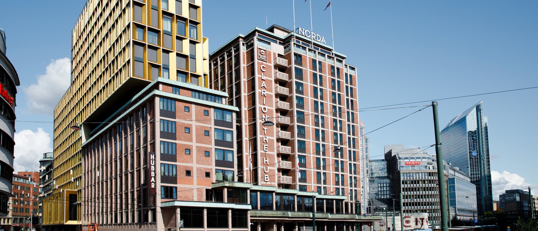 Clarion Hotel The Hub sett fra Arkaden. Med blå himmel i bakgrunnen, travle folk som beveger seg rundt og en trikk som har stoppet foran inngangen. 