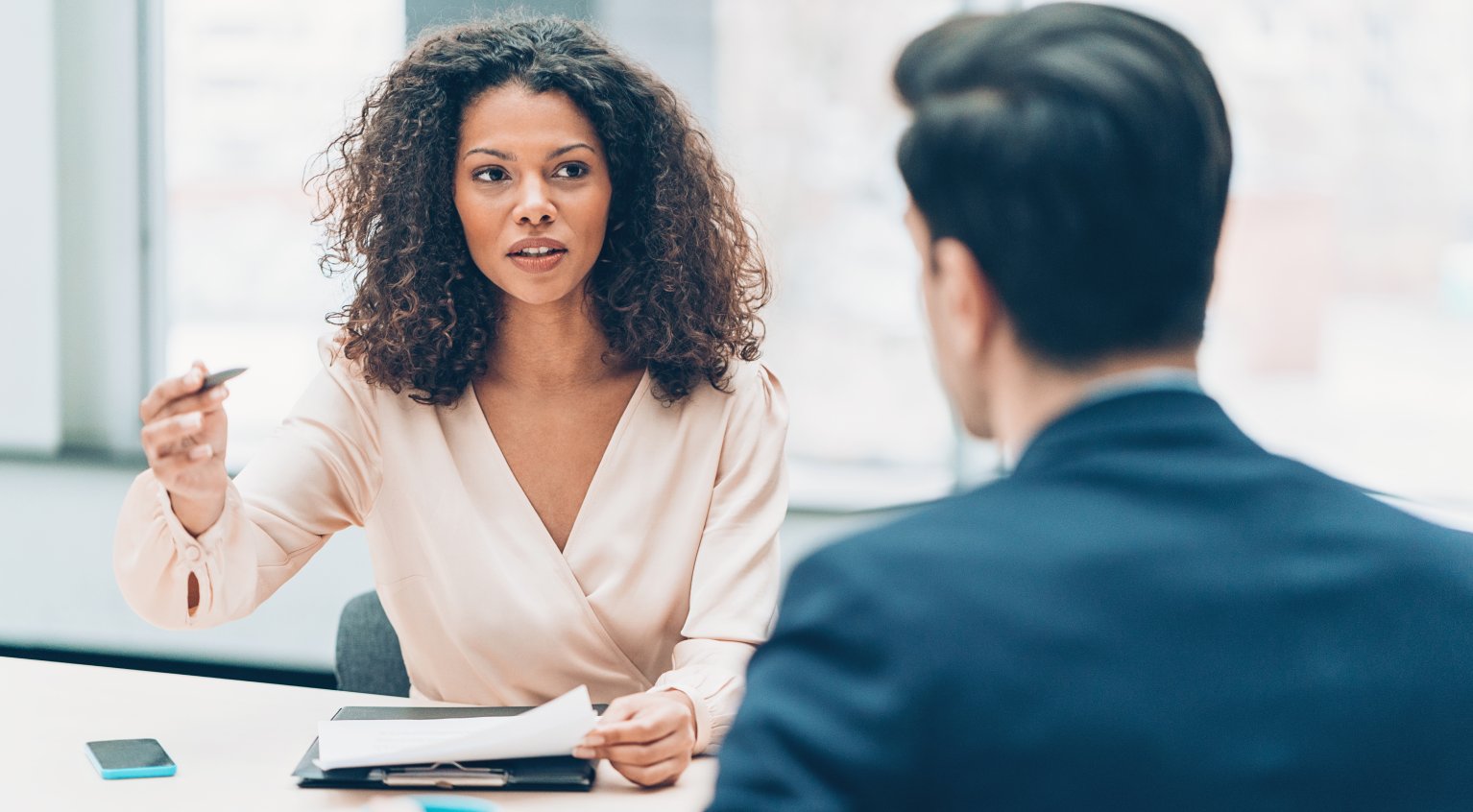 En dame og en mann snakker sammen på et kontor. Mannen sitter med ryggen til og har på seg en blå dress. Damen ser på mannen mens hun holder en penn og har papirer foran seg. Hun har mørkt krøllete hår og lys kjole.